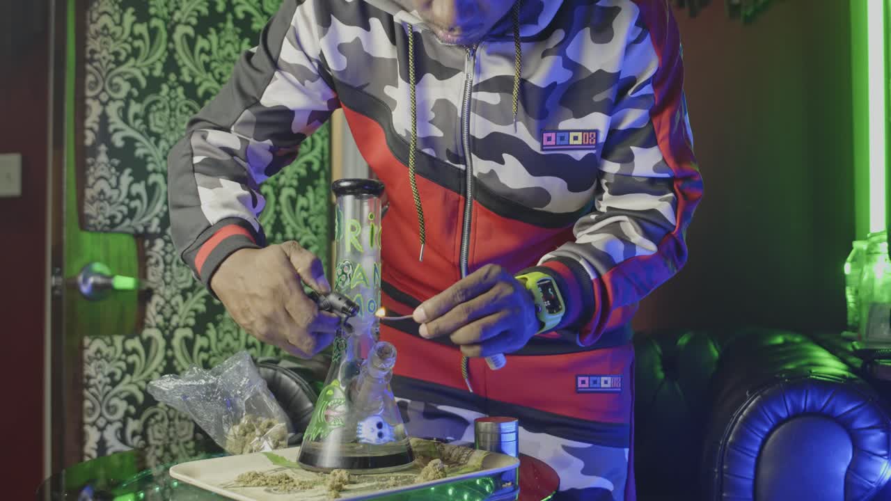 чернокожий человек зажигает фитиль, который он использует, чтобы зажечь марихуанный водяной бонг и берет кусок марихуаны или марихуаны