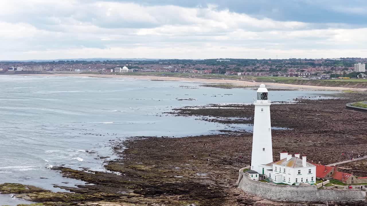Drone camera smoothly approaches St. Mary's Lighthouse on Whitley Bay's rocky shoreline under overcast daylight, highlighting coastal landscape and historic tower