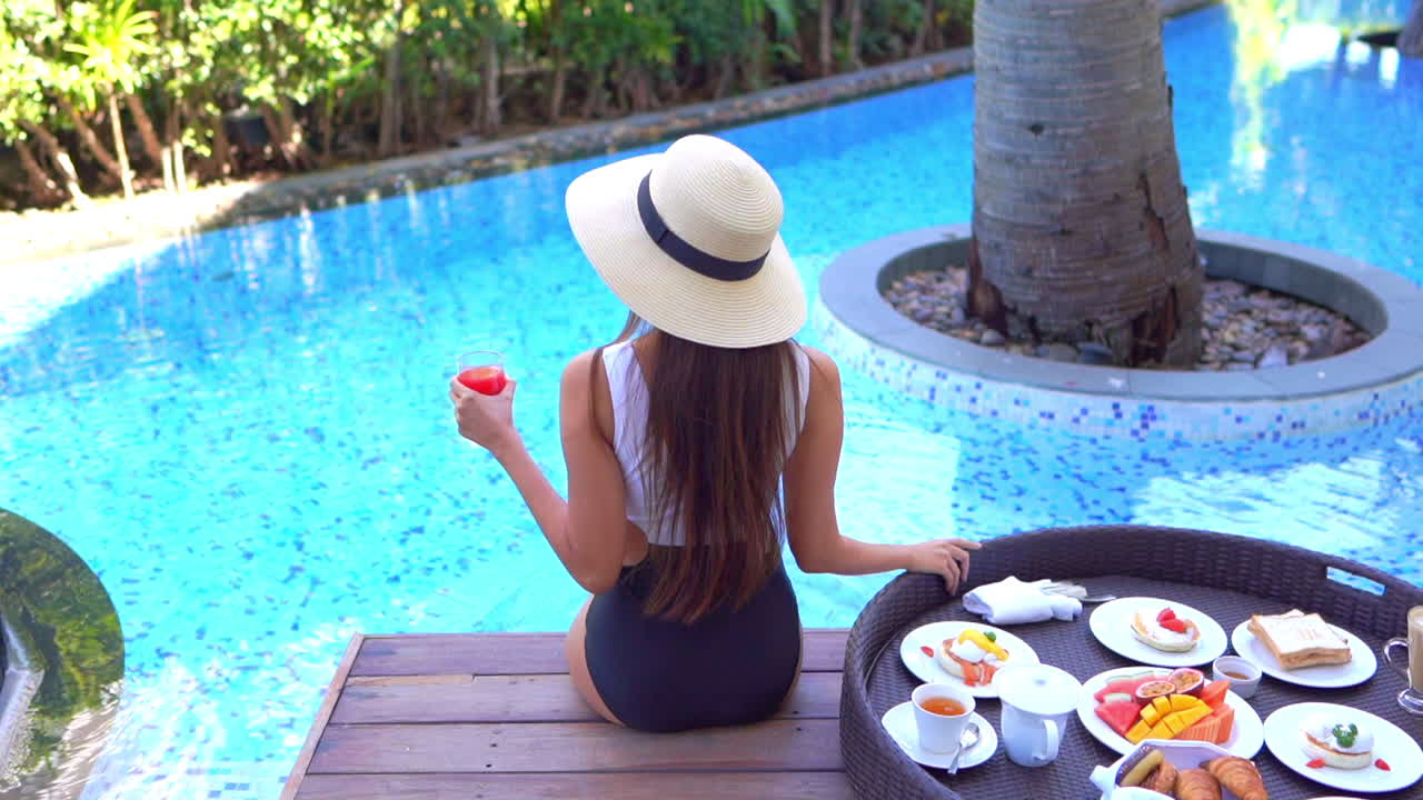 Long haired woman sits by swimming pool with large breakfast tray, back view