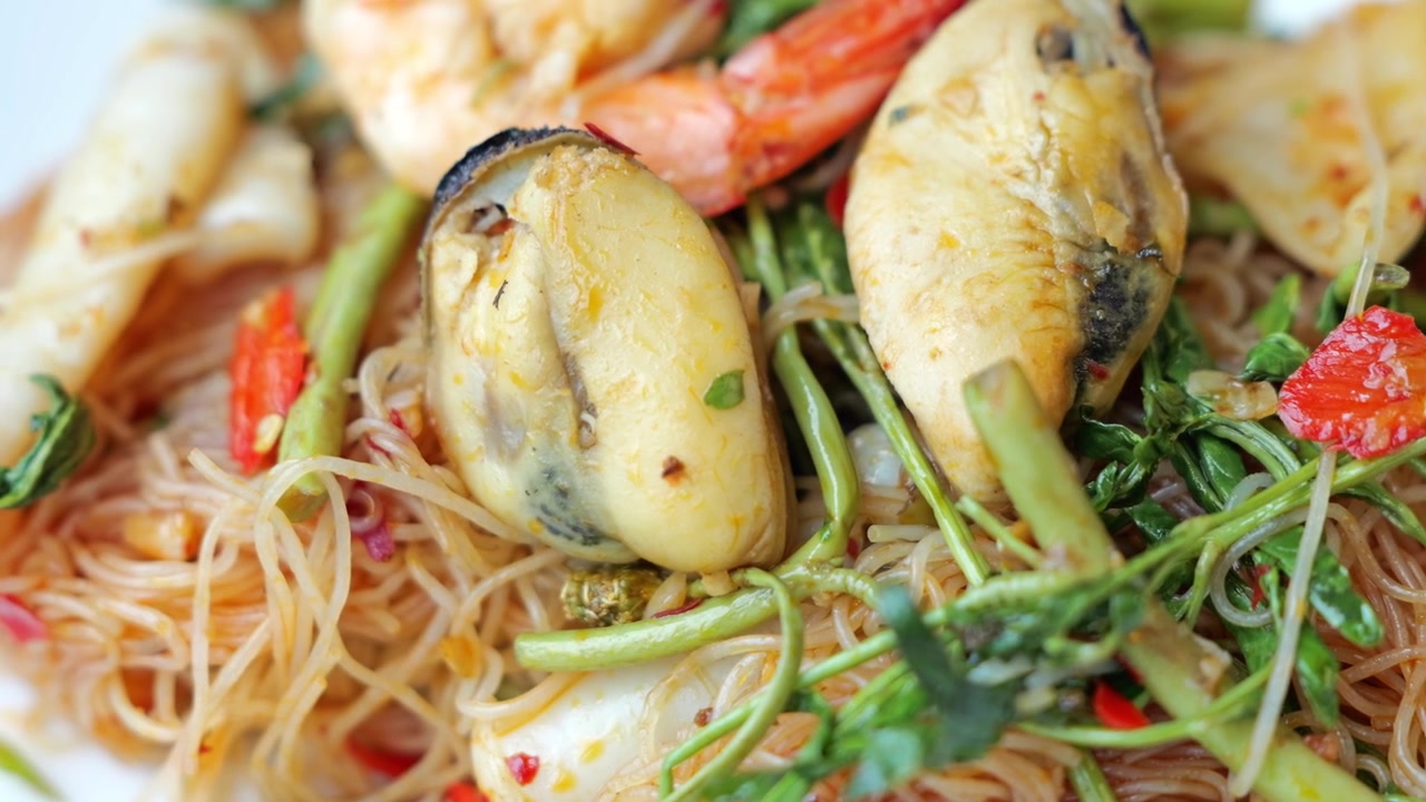 Close-up of spicy stir-fried rice vermicelli with water mimosa, shrimp, squid, and fresh vegetables, served hot with vibrant Thai-style flavors.