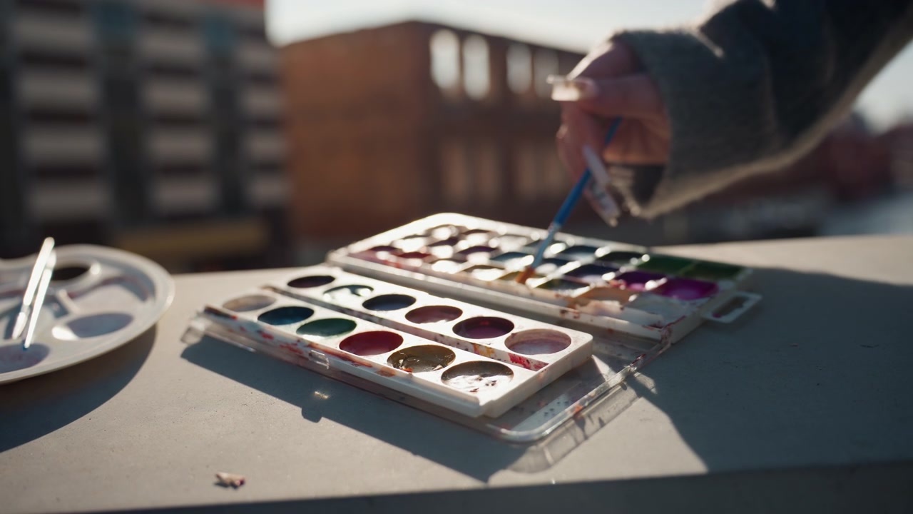 Pretty rooftop watercolor palette and brush, artists hand dips brush into vibrant pans, sunlight warms compact tin on concrete sill, soft bokeh city buildings in background, cozy sweater sleeve