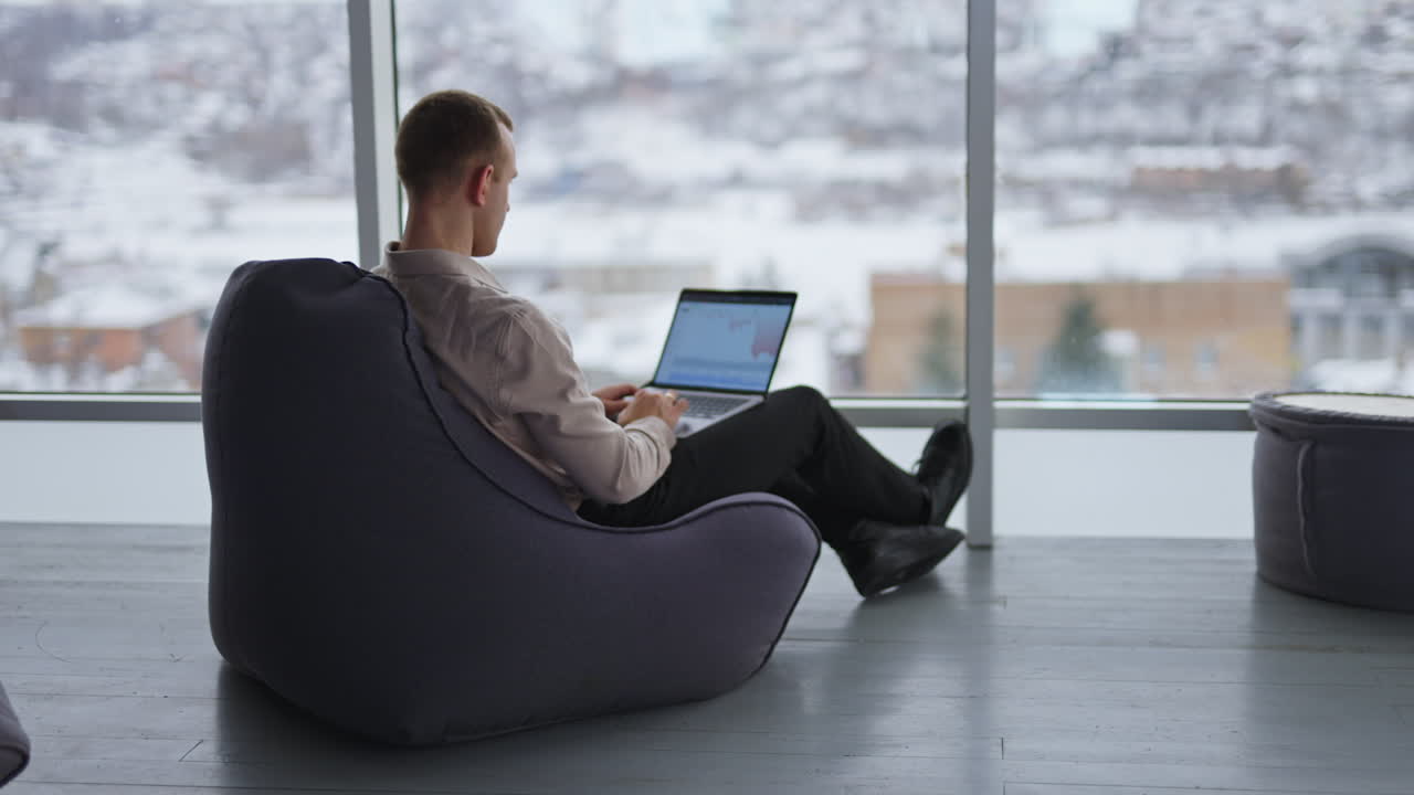 Working man sits with his back to the camera typing on his laptop. Male employee building charts on computer. City panorama in blur at backdrop.