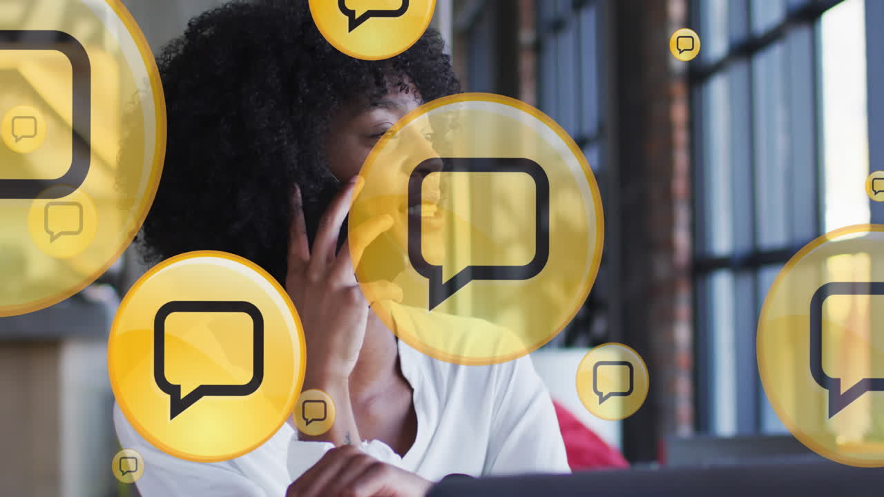 varios iconos de mensajes flotando contra una mujer afroamericana hablando en un teléfono inteligente en la oficina