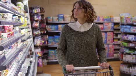 comida, concepto de salud - mujer en un supermercado de pie frente a los estantes del congelador y elige comprar una botella de yogur, tomar uno y ponerlo en el carrito. cámara lenta