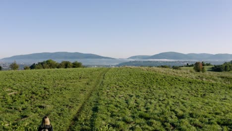 Una-Chica-Rubia-Con-Una-Mochila-Pequeña-Camina-Por-Un-Claro-En-Una-Colina,-En-El-Valle-Hay-Un-Pueblo-Bajo-La-Niebla