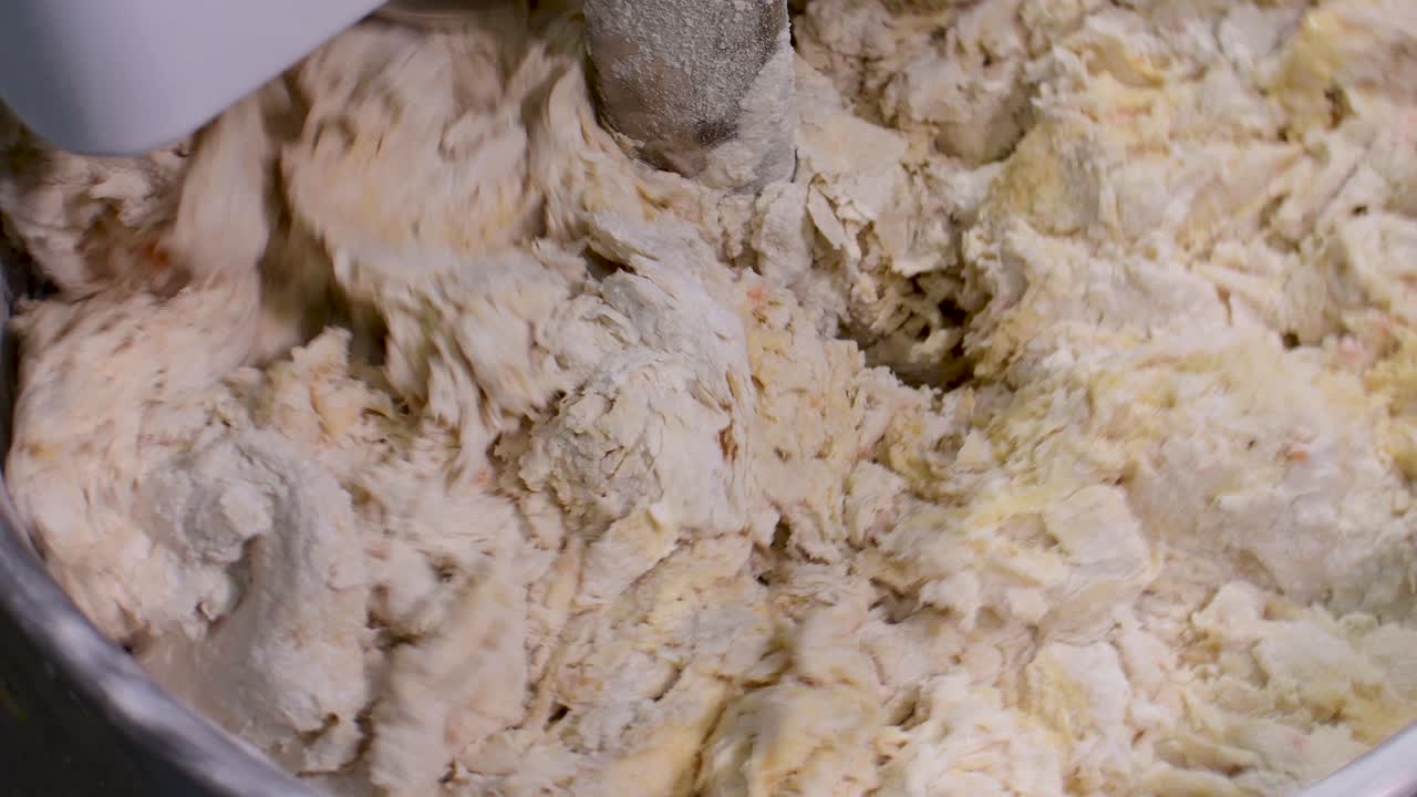 Technical close-up static shot of a professional spiral mixer kneading a buttery yellow brioche dough, capturing the elastic texture and the mechanical process