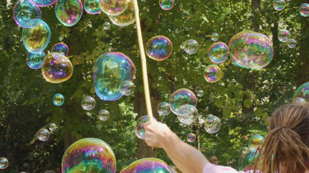 A person uses sticks and string to make large, iridescent soap bubbles outdoors in a sunlit park, surrounded by green trees and natural light