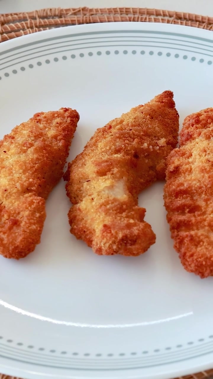 Tongs place golden fried chicken pieces onto a white plate. Bright lighting enhances the crispy texture