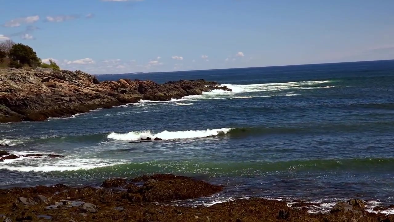 Rocce illuminate dal sole incontrano il mare mosso sotto un cielo blu con nuvole sottili. Nessuna persona, nessun animale. Costa calma, onde forti, terreno accidentato. Perfetto per progetti di viaggio, natura o avventura.