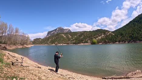 a fisherman in black attire casts a fishing line into a tranquil mountain lake surrounded by lush pine forests and rugged hills under a partly cloudy sky