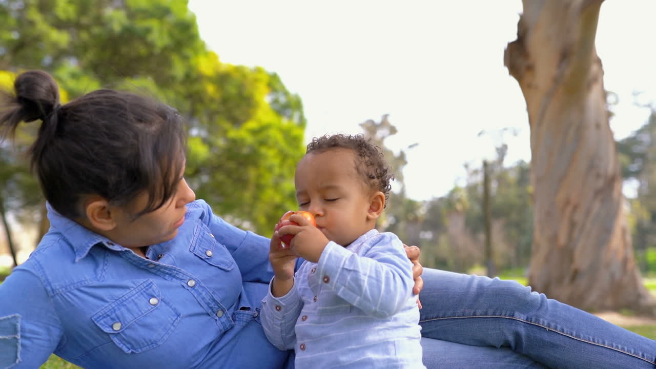 madre acostada en la hierba, mirando a su hijo comiendo manzana, acariciándolo