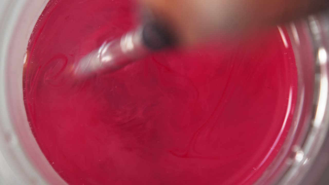 Top down view of red paint releasing and spreading across water from brush swirling