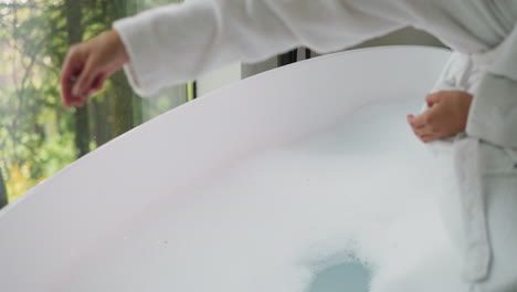 woman pours salt with dry herbs into foamy bathtub closeup. lady in white bathrobe adds aromatic organic product to hot water for bathing in hotel room