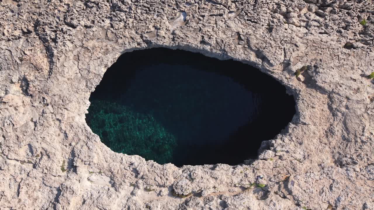 Aerial view of a natural rock formation featuring a large hole that opens to the turquoise sea below. The rugged limestone surface contrasts with the deep blue water, creating a striking coastal scene
