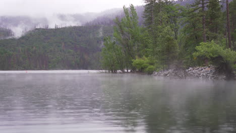 Niebla-Rodando-Sobre-El-Lago-Durante-Una-Lluvia-Ligera-De-Primavera,-Las-Nubes-Cubren-La-Montaña-Boscosa-En-El-Fondo