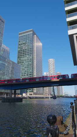 Vertical-Video-Of-Docklands-Light-Railway-Train-Crossing-Bridge-Into-Canary-Wharf-Station-In-Finance-And-Business-District-Of-London-UK