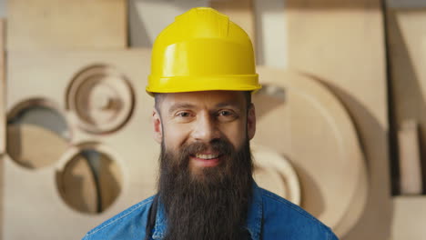 retrato de un hombre barbudo caucásico con casco mirando a la cámara y sonriendo en un taller de carpintería