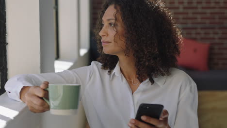 mature-mixed-race-woman-using-smartphone-drinking-coffee-at-home-enjoying-relaxed-morning-browsing-messages-looking-out-window-thinking-contemplative