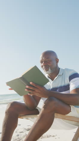 vertical video of senior african american man reading book on deckchair at beach, in slow motion