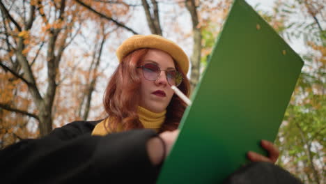 lower angle view of talented mind thoughtfully sketching on canvas, seated with head tilted. immersed in artistic focus, wearing stylish yellow beret and scarf, surrounded by warm autumn color