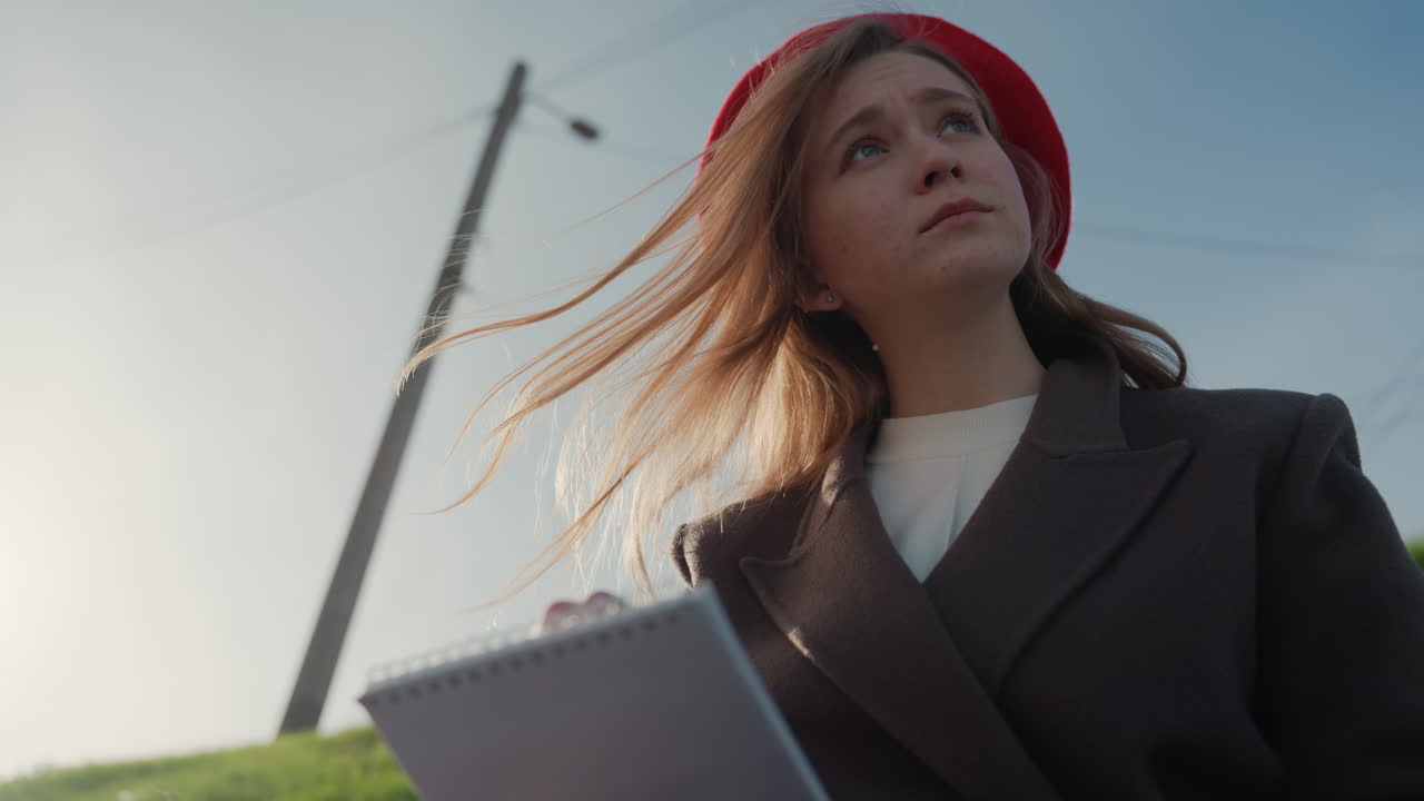 Primo piano di donna bianca che scrive sotto il sole, con berretto rosso e capelli retroilluminati. Composizione cinematografica dal basso, silhouette di un palo del telefono sullo sfondo. Le pagine girano, espressione concentrata e determinata, delicata.