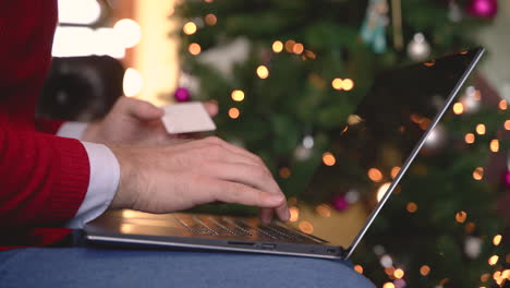 nahaufnahme von mannhänden, die auf einem laptop über ihren beinen tippen, die in der nähe des weihnachtsbaums im wohnzimmer mit weihnachtsdekoration sitzen