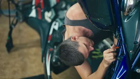 Vertical-video-of-a-man-with-a-short-haircut-in-a-gray-T-shirt-repairing-his-blue-bike-with-a-screwdriver-against-the-background-of-another-motorcycle-in-his-garage-workshop