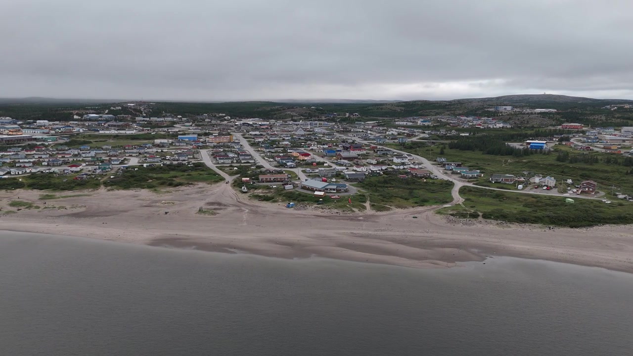 Dronen ilmakuva seuraa vasemmalle Koksoak-joen rantaa Kuujjuaqissa, Nunavikissa, Quebecissä, näyttäen kaupungin, ympäröivän tundramaiseman ja pilvisen taivaan.