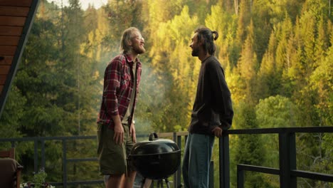 Dos-Amigos-Se-Comunican-Emocionalmente-Y-Se-Ríen-Durante-Un-Picnic-Con-Vistas-A-Un-Bosque-De-Coníferas.-Comunicación-Mientras-Se-Cocina-A-La-Parrilla