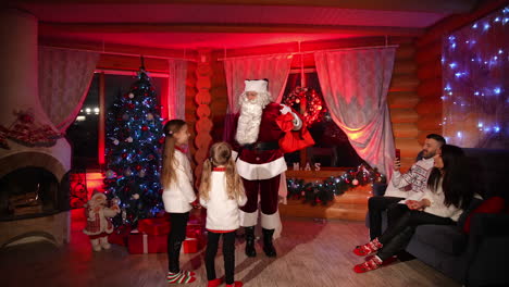 santa claus and happy family indoors. little girls talking to santa near the christmas tree. happy parents sitting on a sofa in decorated room. holidays and celebration.