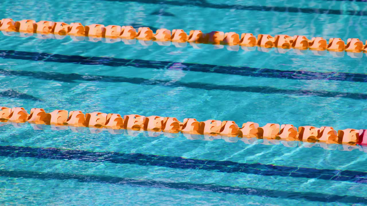 Overhead view of empty swimming pool lanes with clear blue water, orange lane dividers, and sunlight reflections. Subtle water movement, no visible swimmers