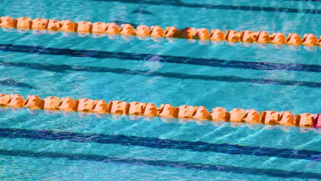 overhead view of empty swimming pool lanes with clear blue water, orange lane dividers, and sunlight reflections. subtle water movement, no visible swimmers