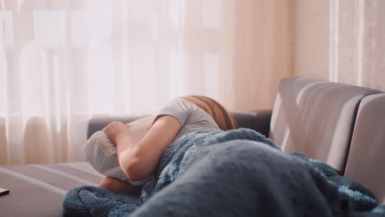 Stressed woman lies curled on sofa with face hidden in pillow under soft sunlight curtain shadows, exhausted from previous workload, unwilling to get up, wrapped in blue blanket as light streams in