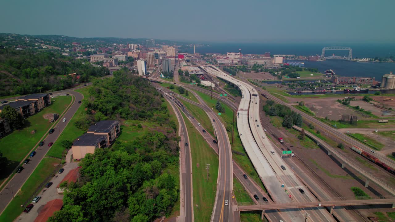 Interstate 35 curving along the hillside toward the Aerial Lift Bridge with rail lines, port industry, and Lake Superior along the Duluth waterfront on a hazy summer day