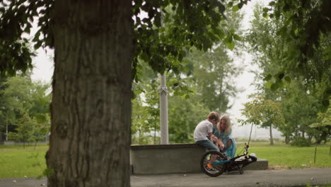 a woman in a blue dress tends to her son's injured leg beside a fallen bicycle in a peaceful park, with a blur view of someone walking in the background