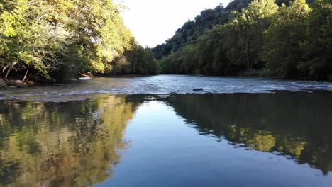 drone flies low over a river with a reflection of the trees on either side of the river