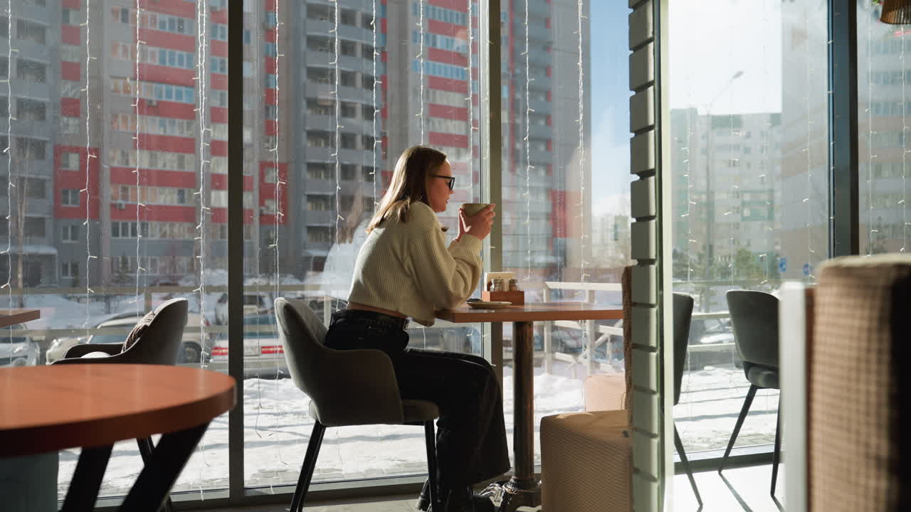 Side view of lecturer seated at cafe table sipping tea near large window, soft sunlight fills cozy space, snowy winter cityscape with parked cars and distant office buildings visible outside