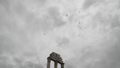 Looking-Up-At-Group-Of-Black-Birds-Circling-High-Above-Stone-Column-Ruin-Against-Moody-Overcast-Clouds