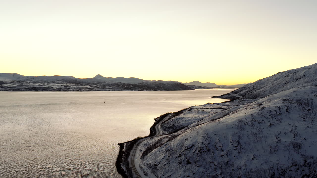 Kinoreife Drohnenaufnahme von verschneiten Bergen und einer kurvenreichen Küstenstraße entlang eines Fjords auf der Insel Ringvassøya nahe Tromsø, Norwegen, bei Sonnenuntergang.
