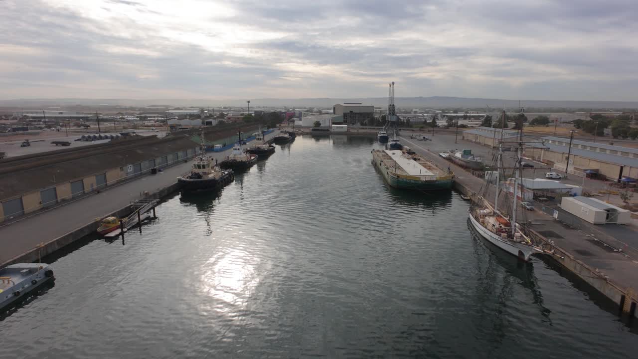 slow flight over port where a number of tugboats are moored by some store houses, there is a old sailboat and a ship amongst the tugboats