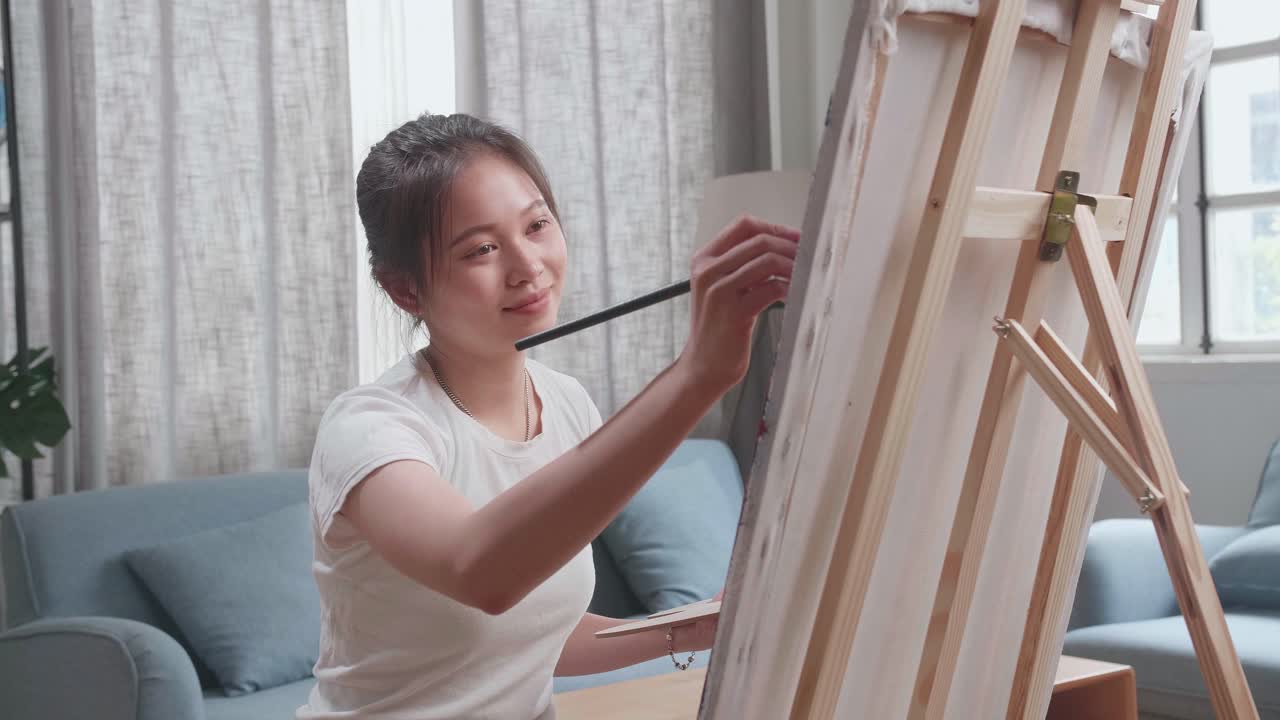 Asian Artist Girl Sitting On Round Wooden Chair Without Backrest Holding Paintbrush And Mixing It With Colour Before Painting On The Canvas