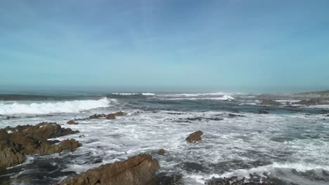 Drone-flying-low-over-boulders-on-a-beach-revealing-breaking-waves,-tides-and-bamboo-as-it-continues-on-to-open-sea