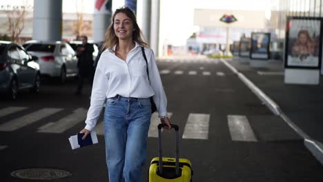 excited, stylish woman walk with trolley yellow case by empty airport terminal outside road, slender female wearing jeans and