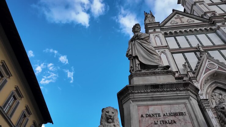 Ripresa dal basso della Basilica di Santa Croce a Firenze, con la statua in marmo di Dante Alighieri di Enrico Pazzi. La facciata gotica, gli allori e un libro evidenziano l'eredità del poeta.