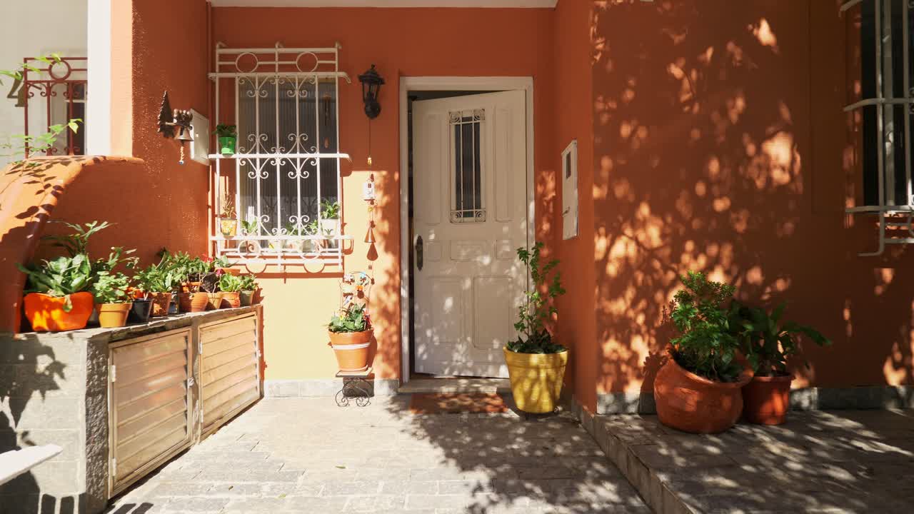A sunny entryway with an open white door, terracotta walls, white window bars, and many potted plants casting leafy shadows across the facade, creating a warm and inviting atmosphere.