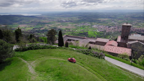 aerial pan of red car on green hill by town of cortona in tuscany