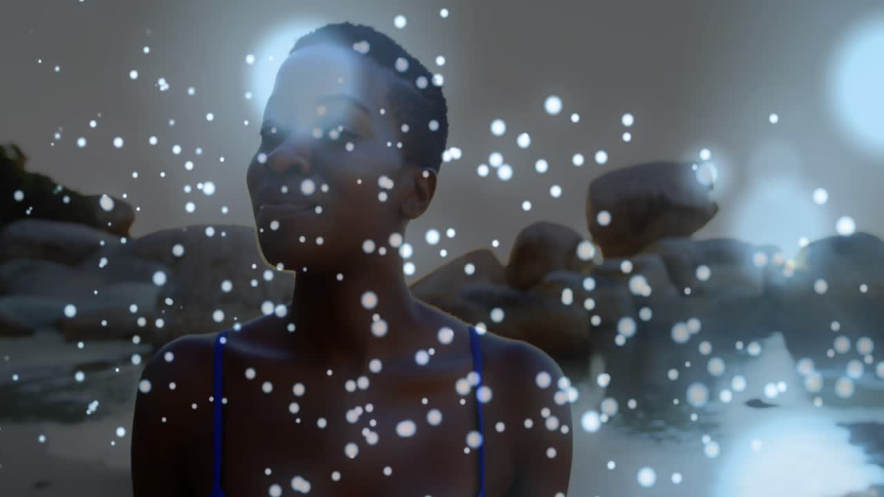 Glowing blue spots of light against african american woman walking on the rocks near the sea
