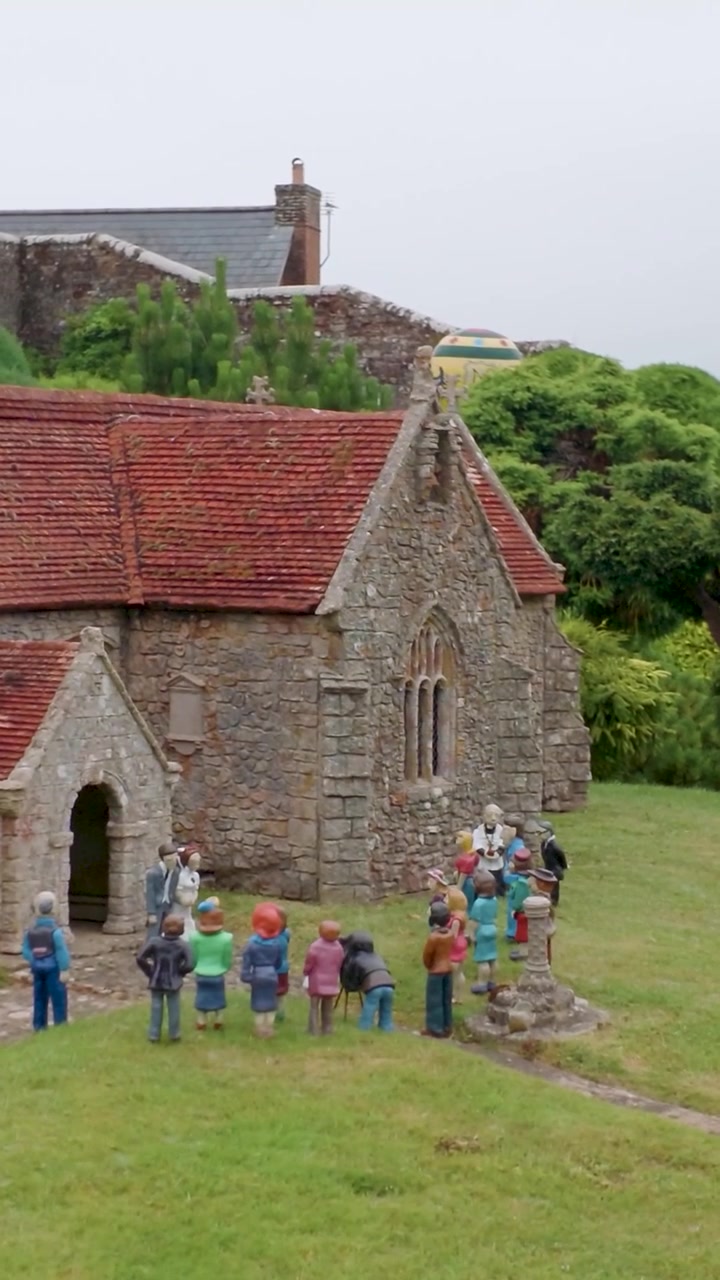 Model af All Saints Church, Godshill, Isle of Wight med miniature bryllupsgæster samlet udenfor, mens kameraet panorerer ned og til højre, delvist afsløret bag et træ i en landsbyhaveudstilling. Lodret