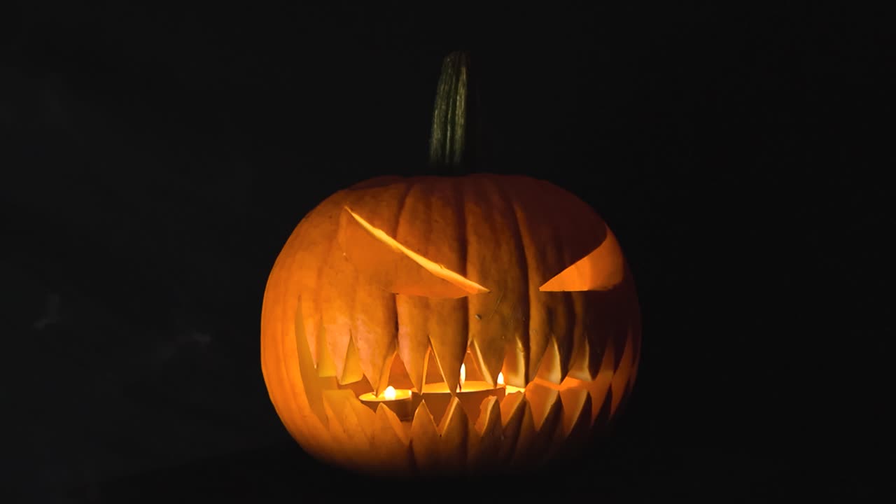 Carved Halloween jack o lantern pumpkin that has an evil face carved into it where candles burn and create smoke in the foreground in front of a black studio background