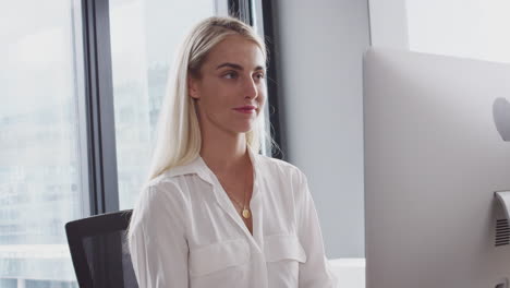 Millennial-Asian-woman-sitting-at-an-office-desk-using-a-computer-and-drinking-coffee,-waist-up